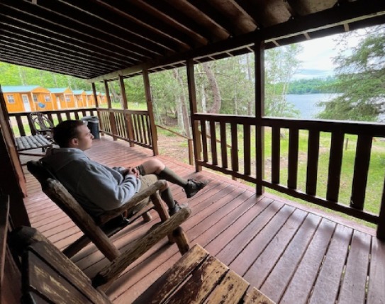 A man sits in a wooden chair on a clean wooden deck, looking out over a green trail that leads down to Lake Killian